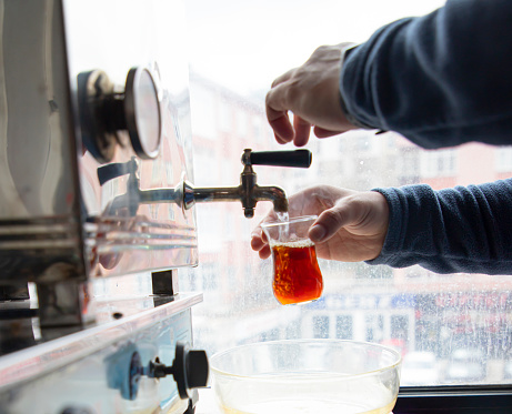 pouring tea from a big teapot into a teacup. steel chai boiler for public spaces. selective focus on red glass. Tea time at office