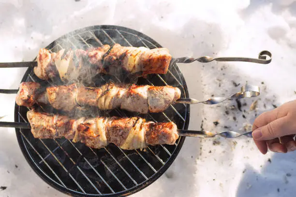 Photo of BBQ shashlik in the snow in winte. Kebabs in nature. Female hand turns skewers with meat