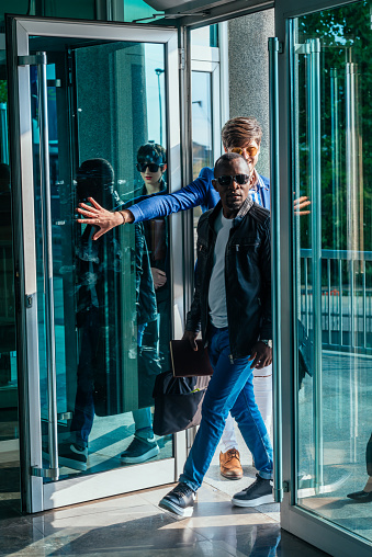 Two multinational young businessmen entering an office building with glass doors.