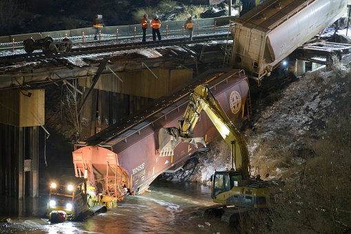On February 12, 2022 in Denver, Colorado, several railroad cars of a BNSF Railway train derailed from their tracks on a bridge Saturday morning and fell into the South Platte River, blocking a riverside trail. At night, workers use heavy equipment to pull the stranded cars out of the river.