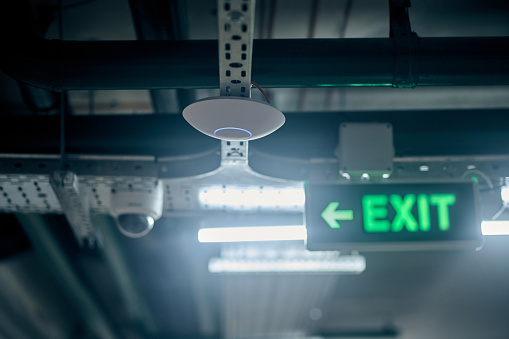 Optical smoke detector mounted on the ceiling beside the illuminated exit sign with a green arrow and the surveillance camera