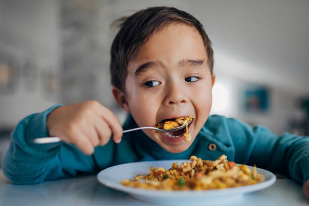 A front view of a hungry mixed-race boy eating fried rice for lunch with a spoon. The background is blurred. Portrait of a beautiful three-year-old boy, who enjoys eating Thai cuisine prepared by his mother for lunch. eating fried rice stock pictures, royalty-free photos & images