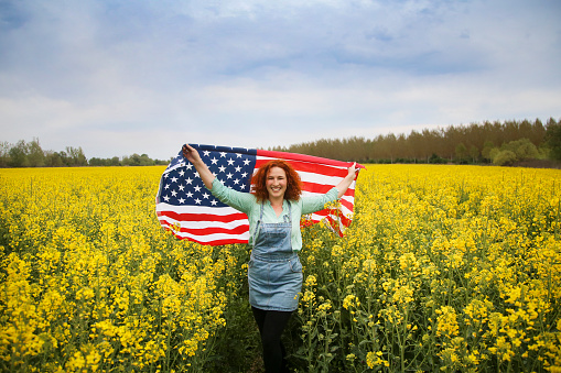 Happy smiling young woman with American flag posing in an oilseed or canola field. About 25 years, female Caucasian redhead.