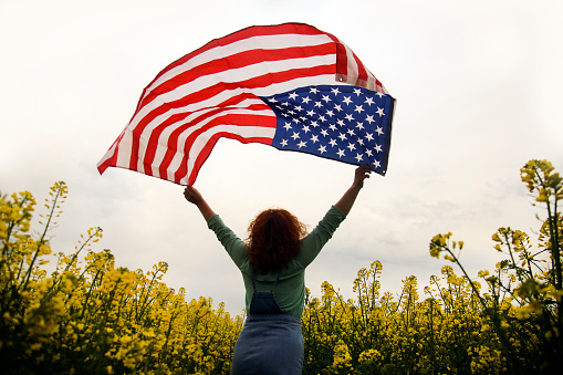 Happy smiling young woman with American flag posing in an oilseed or canola field. About 25 years, unrecognizable female Caucasian redhead.