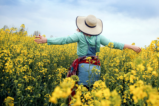 Happy smiling young woman with outstretched arms posing in an oilseed or canola field. About 25 years, unrecognizable female Caucasian redhead.