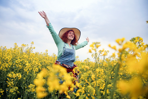 Happy smiling young woman with outstretched arms posing in an oilseed or canola field. About 25 years, female Caucasian redhead.