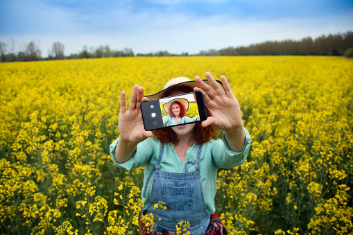 Happy smiling young woman taking a selfie in an oilseed or canola field. About 25 years, female Caucasian redhead.