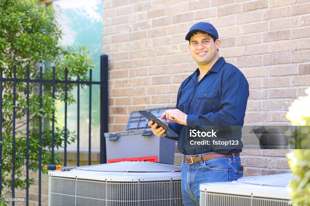 Latin descent young man, blue collar air conditioner repairman at work. Latin descent blue collar air conditioner repairman working at residential home.  He prepares to begin work by gathering appropriate tools and referring to digital tablet. Air Conditioner Stock Photo