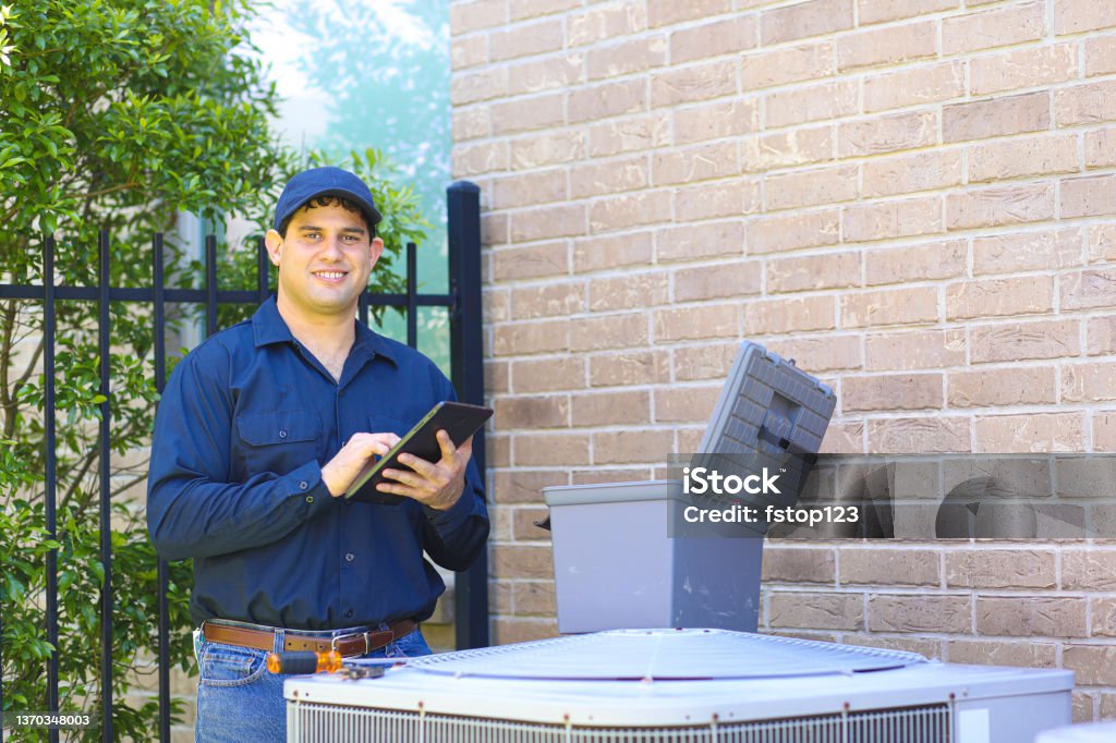 Latin descent young man, blue collar air conditioner repairman at work. Latin descent blue collar air conditioner repairman working at residential home.  He prepares to begin work by gathering appropriate tools and referring to digital tablet. Air Conditioner Stock Photo