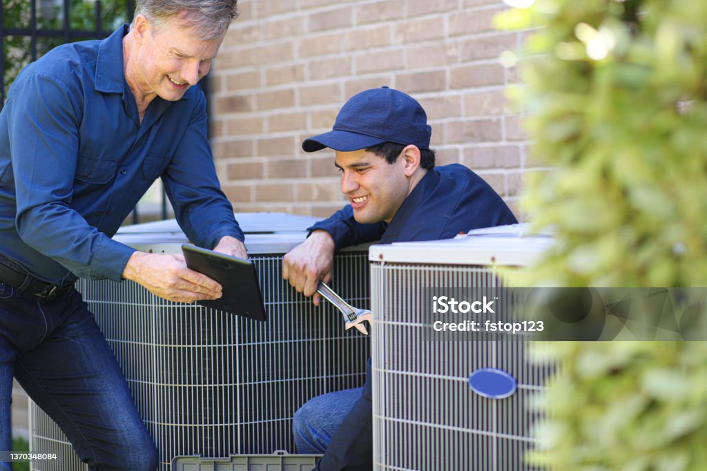 Multi-ethnic team of blue collar air conditioner repairmen at work. Multi-ethnic, mixed age team of blue collar air conditioner repairmen working at residential home.  They prepare to begin work by gathering appropriate tools and referring to digital tablet. Air Conditioner Stock Photo