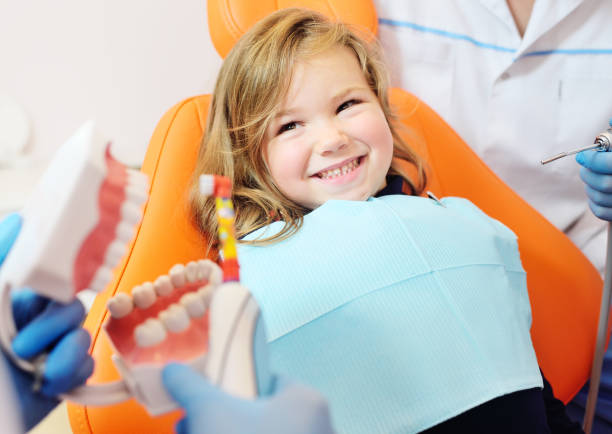 pediatric dentist shows how to properly brush her teeth to a little girl who is sitting in an orange dental chair and smiling stock photo