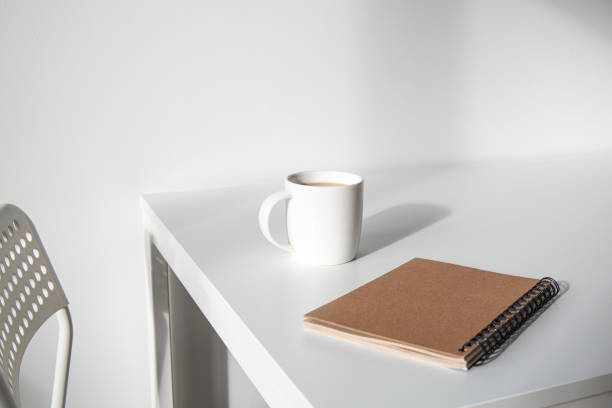 istock White cup of coffee, a notebook on a white table, there is a chair next to it. 1370357204