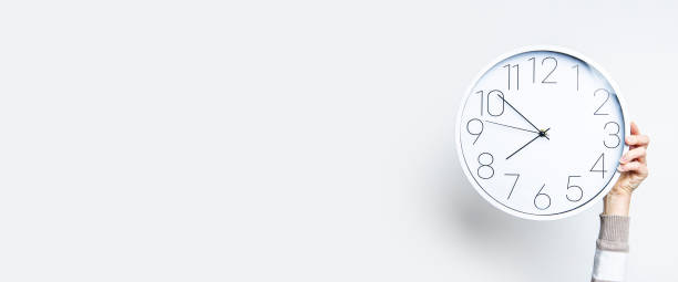 istock Woman's hand holds a large white clock against a light wall. Banner. 1370358419