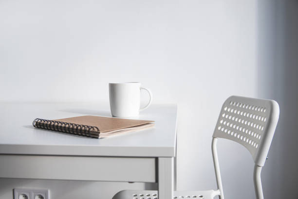 istock White cup of coffee, a notebook on a white table, there is a chair next to it. 1370358477