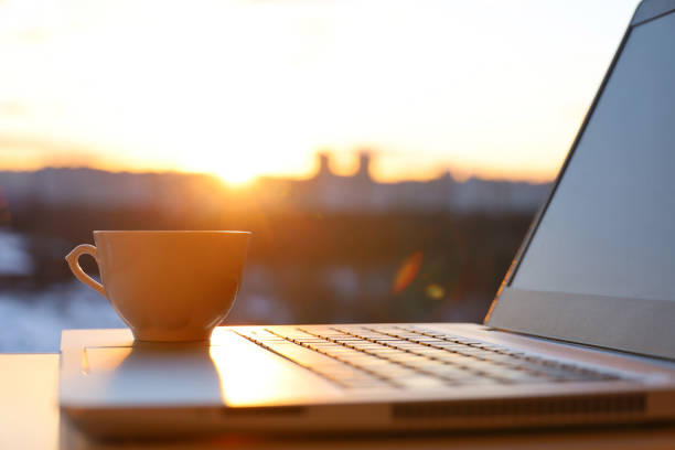 tasse de café sur ordinateur portable contre la fenêtre avec soleil - in the evening photos et images de collection