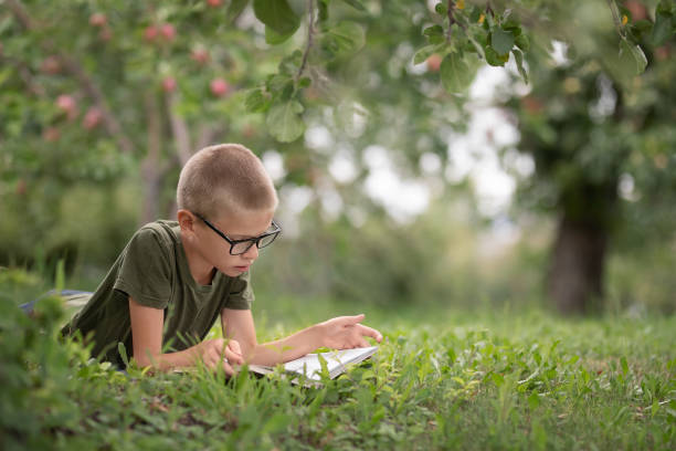 a boy with glasses lies on the grass in the park and reads a book