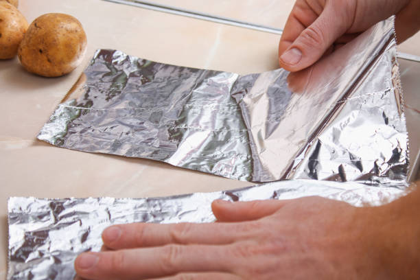 un hombre arranca el papel de aluminio del dispensador de plástico para envolver las papas en él y hornearlas en el horno. - rolled up foil paper gray fotografías e imágenes de stock