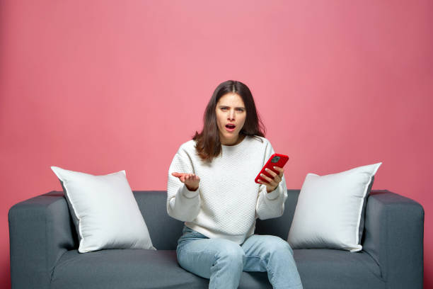 Shocked annoyed young woman holds phone irritated by by bad news or spam in message online, sitting on sofa Shocked annoyed young woman holds phone irritated by by bad news or spam in message online, sitting on sofa. High quality photo phone spam photos stock pictures, royalty-free photos & images