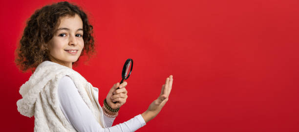 one small caucasian girl ten years old with curly hair front view portrait close up standing in front of red background looking to the camera holding magnifying lens education and learning concept - magnifying glass lens holding europe imagens e fotografias de stock