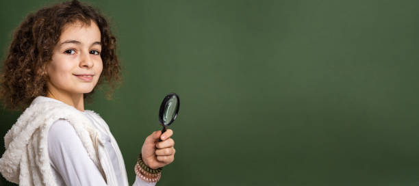 one small caucasian girl ten years old with curly hair front view portrait close up standing in front of green background looking to the camera holding magnifying lens education and learning concept - magnifying glass lens holding europe imagens e fotografias de stock