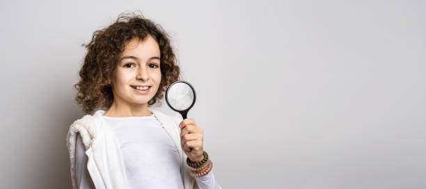 one small caucasian girl ten years old with curly hair front view portrait close up standing in front of white background looking to the camera holding magnifying lens education and learning concept - magnifying glass lens holding europe imagens e fotografias de stock