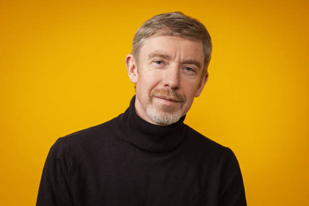 Close-up studio portrait of a 50 year old bearded man in a blue sweater on a yellow background stock photo
