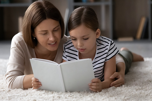 Close up smiling mother with little girl reading book together, lying on warm floor at home, hugging, loving mom and adorable daughter kid learning, enjoying leisure time, educational activity