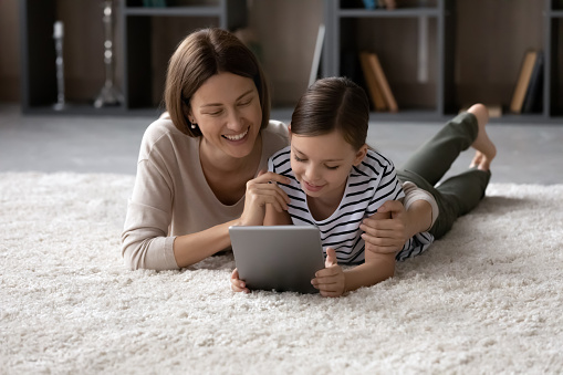Smiling mother with daughter hugging, using computer tablet together, lying on warm floor at home, happy mom with little girl kid having fun with modern device, chatting online, watching video