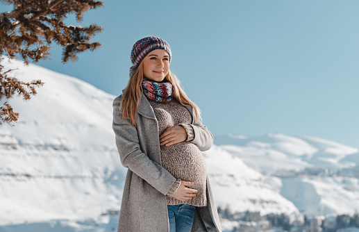 Nice Pregnant Woman With Pleasure Touching her Belly. Enjoying Bright Sun Light in Snowy Mountains. Spending Pregnancy in Winter Mountainous Resort.