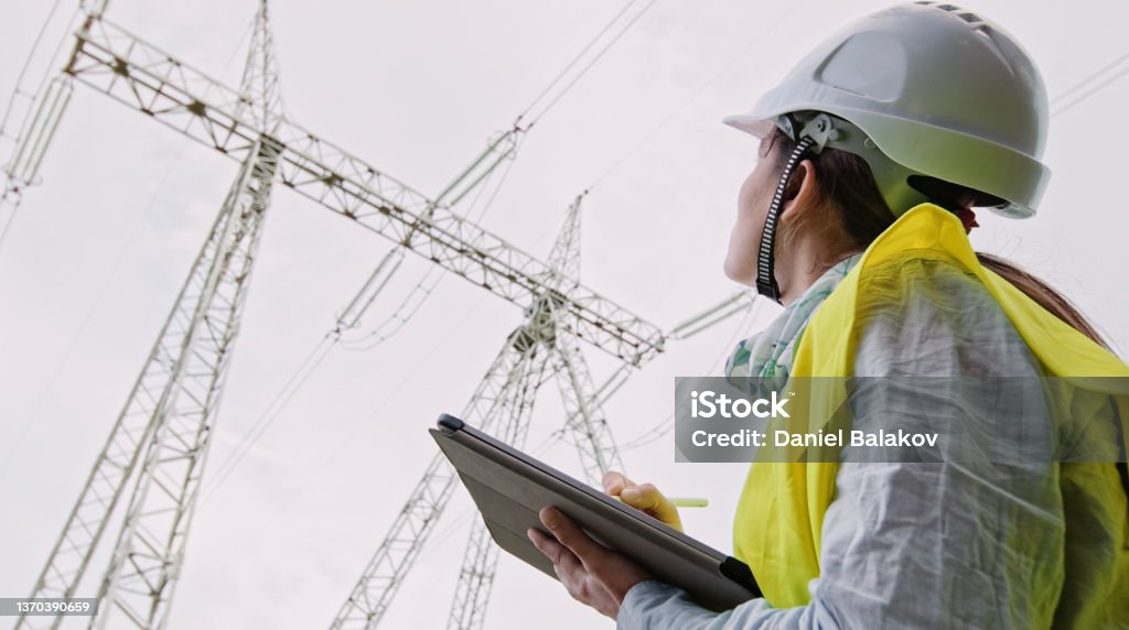 High voltage female engineer working on the field. Female electrical engineer working on Electrical Pylons Translation, checking the condition of the Electrical Power Pole components. Electricity Substation Stock Photo