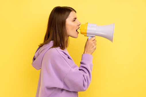 Side view portrait of young adult beautiful woman screaming in megaphone and announcing discounts in mall, wearing purple hoodie. Indoor studio shot isolated on yellow background.
