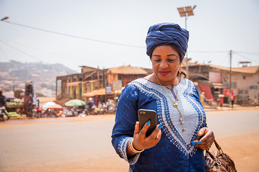 An African woman dressed in a traditional dress looks at the screen of her smartphone. 50-year-old woman in an African village uses technology