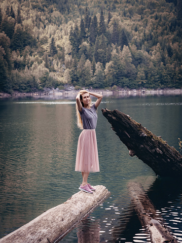 A long-haired woman stands on a log lying in a forest lake near a large trunk of broken tree sticking out of the water. National Nature Reserve