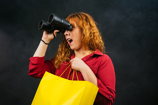 surprised caucasian woman in a red dress with binoculars and a shopping bag on a black background with fog