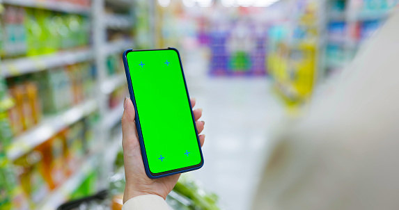 close up of asian woman holds smartphone with green screen and shopping cart in cleaning supplies aisle at grocery store