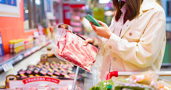 close up of asian young woman wear face mask is shopping for meat and scanning barcode with smart phone at supermarket