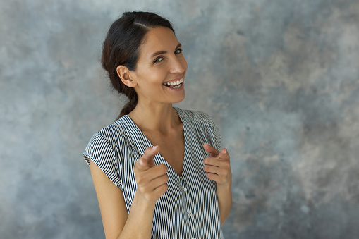Cheerful slim woman of middle age in striped trendy clothes, laughing after getting the joke, giving mocking gesture at camera with index fingers, isolated on background with copy space for your text