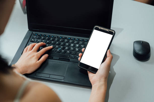 istock Top view mockup image of young woman hands holding blank mobile phone while using laptop with blank black desktop screen on table 1370421187