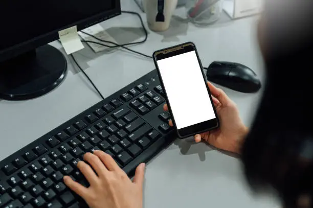 Photo of Top view mockup image of woman hands holding blank mobile phone while using computer with blank black desktop screen on table