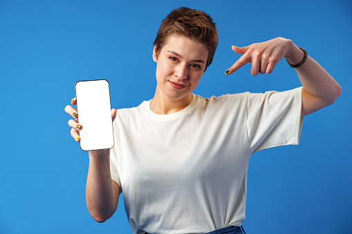Portrait of a young woman showing blank screen mobile phone while standing over blue background, close up