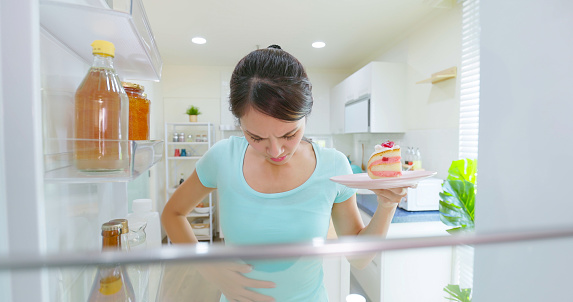 asian woman intending to lose weight so she can not eat High calorie food - girl takes cake from opened refrigerator and worried about her weight in kitchen at home