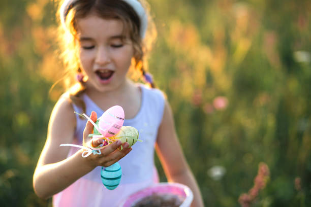 jolie fille drôle avec des œufs de pâques peints au printemps dans la nature dans un champ avec la lumière dorée du soleil et des fleurs. vacances de pâques, lapin de pâques avec oreilles, œufs colorés dans un panier. mode de vie - easter or easter bunny or easter egg or easter basket not business not silhouette photos et images de collection