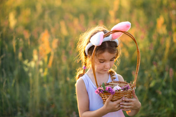 jolie fille drôle avec des œufs de pâques peints au printemps dans la nature dans un champ avec la lumière dorée du soleil et des fleurs. vacances de pâques, lapin de pâques avec des oreilles, œufs colorés dans un panier. mode de vie - easter or easter bunny or easter egg or easter basket not business not silhouette photos et images de collection