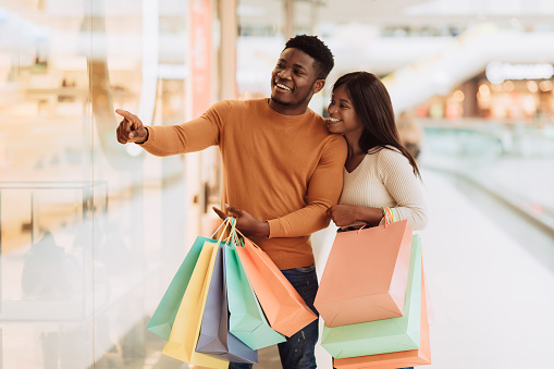 Great Offer. Happy Black Man Shopping In Mall With Excited Girlfriend, Pointing At New Clothing In Showcase, Showing Good Price Discount, Cheerful Young African American Couple Enjoying Seasonal Sales