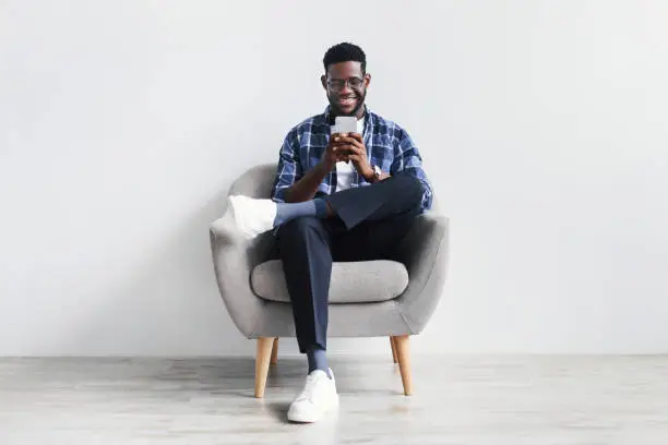 Handsome young black guy using cellphone, surfing web or social media, sitting in armchair, enjoying modern technologies, checking new mobile app against white studio wall, full length