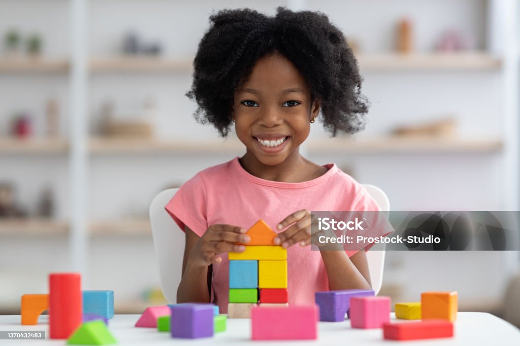 Happy little black girl playing with colorful wooden blocks Happy little african american girl with bushy hair playing with colorful wooden blocks and smiling at camera, enjoying table games while visiting psychologist or playing alone at home, closeup Puzzle Cube Stock Photo