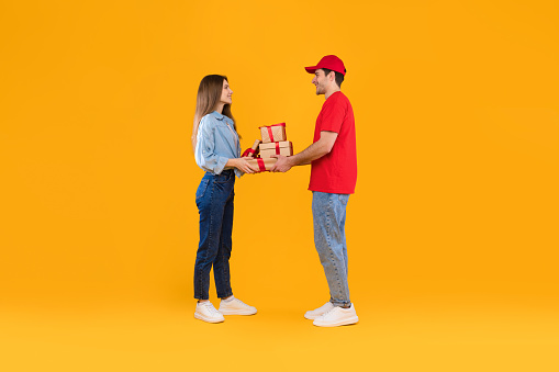 Courier Guy Giving Gifts To Customer Woman Delivering Presents Standing Over Yellow Studio Background. Deliveryman And Female Holding Wrapped Cardboard Boxes. Delivery Service Concept. Full Length