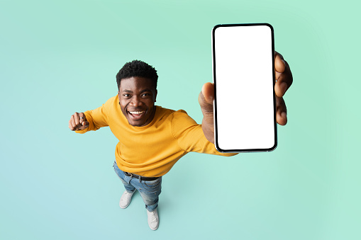 Chico negro emocionado que demuestra el teléfono inteligente con pantalla blanca en blanco, que muestra espacio de copia gratuito para su anuncio, maqueta photo