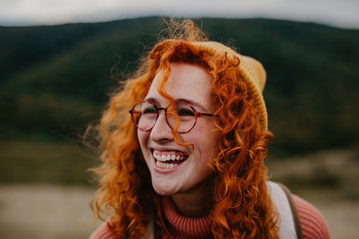 Spontaneous close up image of a cute red hair girl with eyeglasses on enjoying outdoors on a beautiful sunny and windy day, facing the landscape and smiling. Radiating happiness. She's urban dressed, having a peach colored turtleneck and an ocher cap with a backpack