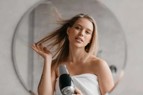 Haircare. Young woman drying and styling hair with hairdryer, making hairstyle in modern bathroom interior. Happy lady using hair dryer enjoying beauty routine after morning shower